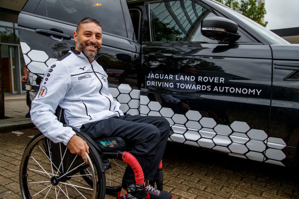 Mann lacht und sitzt im Rollstuhl neben Jaguar Land Rover Driving Towards Fahrzeug