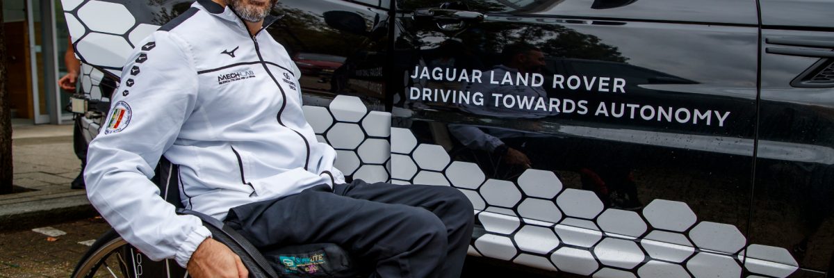 Mann lacht und sitzt im Rollstuhl neben Jaguar Land Rover Driving Towards Fahrzeug