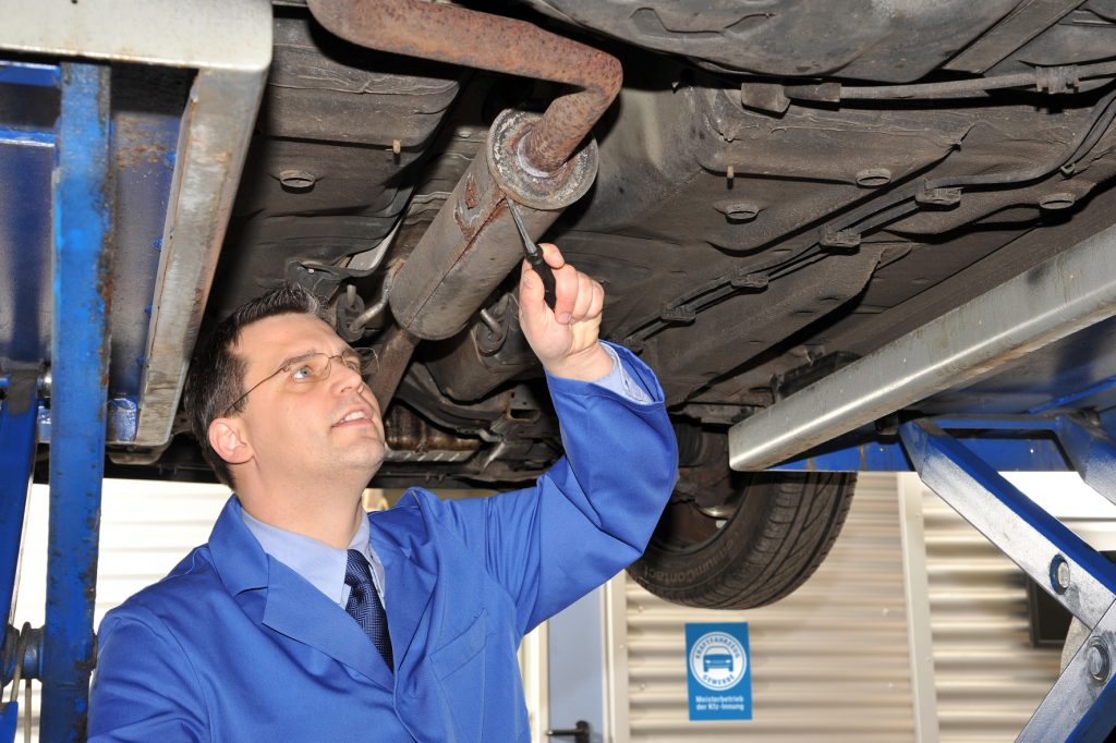 Mann unter Hebebühne in der Werkstatt mit Schraubenzieher am Auspuff.