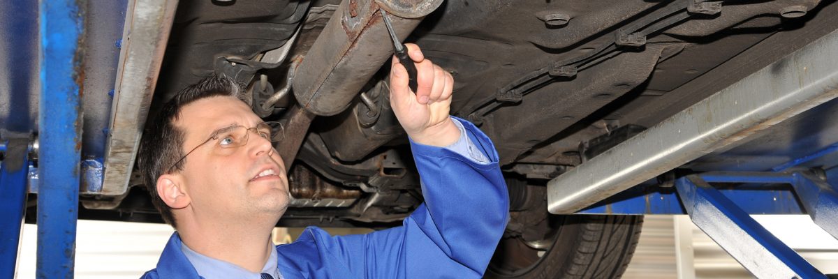 Mann unter Hebebühne in der Werkstatt mit Schraubenzieher am Auspuff.