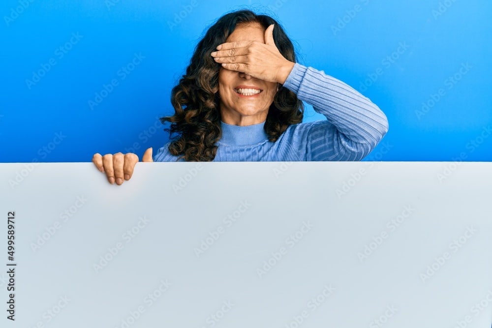 Middle age hispanic woman holding blank empty banner smiling and laughing with hand on face covering eyes for surprise. blind concept.
